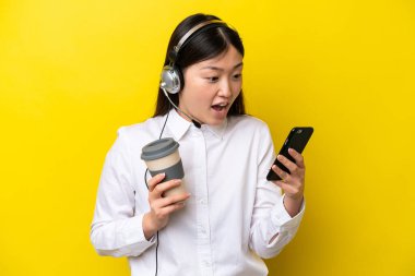Tele pazarlamacı Çinli bir kadın sarı arka planda izole edilmiş bir kulaklıkla çalışıyor. Elinde kahve ve cep telefonu var.
