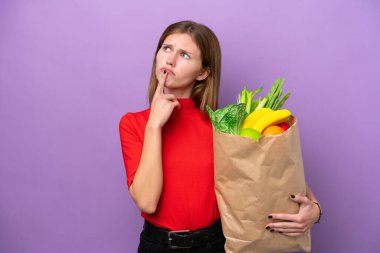 Mor arka planda izole edilmiş alışveriş torbası tutan genç bir İngiliz kadın yukarı bakarken şüphe duyuyor.