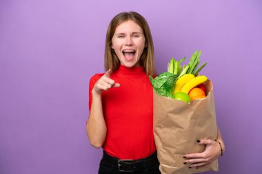 Mor arka planda izole edilmiş alışveriş torbası tutan genç İngiliz kadın şaşırmış ve ön tarafı işaret ediyor.