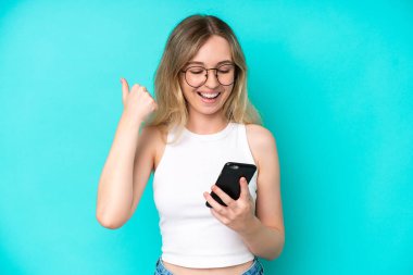 Blonde English young girl isolated on blue background using mobile phone and doing victory gesture