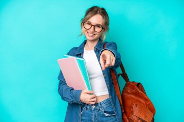 Genç bayan öğrenci mavi arka planda izole edilmiş, mutlu bir ifadeyle ön tarafı işaret ediyor.