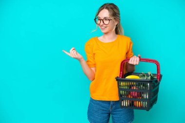 Sarışın İngiliz genç kız elinde yiyecek dolu bir sepet tutuyor mavi arka planda izole edilmiş bir şekilde parmak ucunda bir ürün sunuyor.