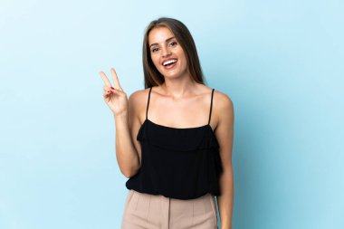 Mavi arka planda izole edilmiş genç Uruguaylı kadın gülümsüyor ve zafer işareti gösteriyor.