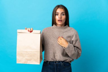 Bir paket yemek alan bir kadın mavi arka planda izole edilmiş, şaşırmış bir yüz ifadesi ile