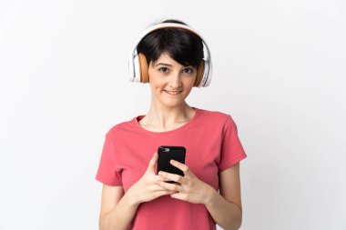 Kısa saçlı bir kadın beyaz arka planda izole edilmiş. Cep telefonuyla müzik dinliyor ve ön tarafa bakıyor.