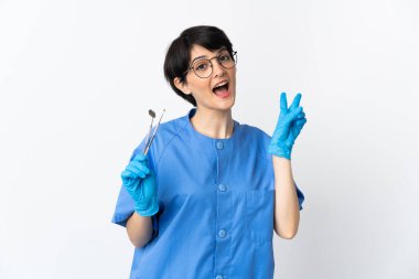 Beyaz arka planda izole edilmiş aletleri tutan dişçi kadın gülümsüyor ve zafer işareti gösteriyor