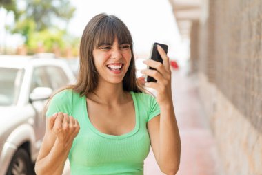 Dışarıda cep telefonu kullanan ve zafer jesti yapan genç bir kadın.