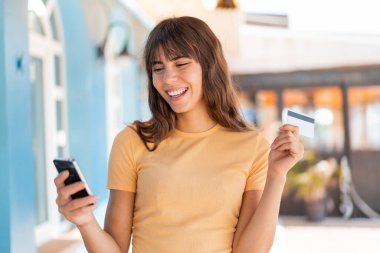 Dışarıda kredi kartıyla cep telefonuyla alışveriş yapan genç bir kadın.