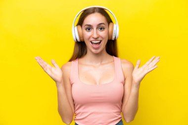 Sarı arka planda izole edilmiş genç beyaz kadın şaşırdı ve müzik dinliyor.