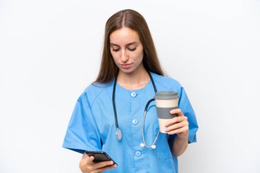 Young nurse woman over isolated on white background holding coffee to take away and a mobile