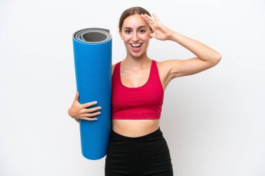 Beyaz arka planda, sürpriz bir ifadeyle izole edilmiş bir minderi tutarken yoga derslerine giden beyaz, beyaz bir kadın.