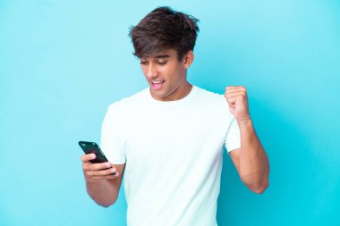 Genç beyaz adam mavi arka planda izole edilmiş cep telefonu kullanıyor ve zafer jesti yapıyor.