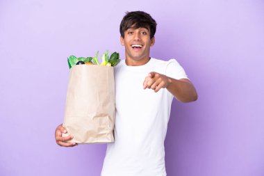 Elinde bir alışveriş çantası tutan genç adam mor arka planda izole edilmiş şaşırmış ve ön tarafı işaret ediyor.