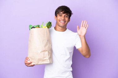 Elinde alışveriş çantası tutan genç bir adam mor arka planda izole edilmiş mutlu bir ifadeyle selamlıyor.