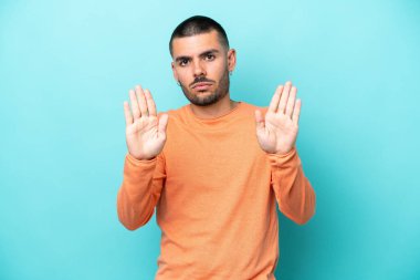 Mavi arka planda izole edilmiş genç beyaz adam dur hareketi yapıyor ve hayal kırıklığına uğruyor.