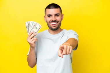 Kafkasyalı genç adam sarı arka planda izole edilmiş bir sürü para alıyor şaşırmış ve ön tarafı işaret ediyor.