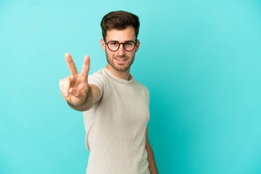 Mavi arka planda izole edilmiş genç beyaz adam gülümsüyor ve zafer işareti gösteriyor.