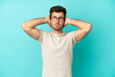 Kafkasyalı yakışıklı adam mavi arka planda izole edilmiş hayal kırıklığına uğramış ve kulaklarını kapatmış.