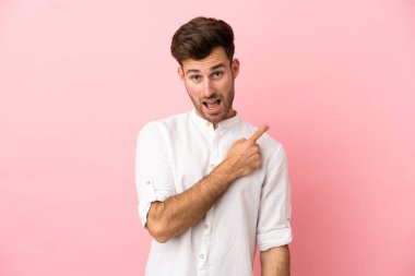 Young caucasian handsome man isolated on pink background surprised and pointing side