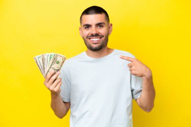 Kafkasyalı genç adam, sarı arka planda izole edilmiş bir şekilde bir sürü para alıyor.