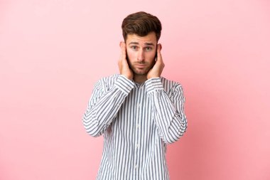 Pembe arka planda izole edilmiş genç beyaz yakışıklı adam hayal kırıklığına uğramış ve kulaklarını kapatmış.