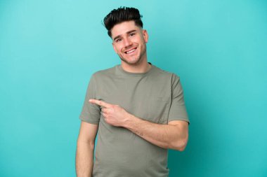 Mavi arka planda izole edilmiş genç beyaz adam şaşırmış ve işaret ediyor.