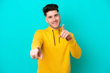 Mavi arka planda izole edilmiş genç beyaz adam mutlu bir ifadeyle ön tarafı işaret ediyor.
