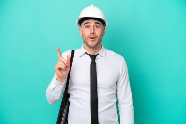 Miğferli ve elinde mavi arka planda izole edilmiş planları olan genç bir beyaz adam çözümü fark ederken parmağını kaldırmayı amaçlıyor.