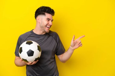 Yakışıklı genç futbolcu sarı arka planda izole edilmiş, parmağını yana doğru gösteren ve bir ürün sunan.