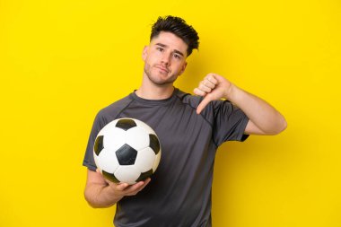 Yakışıklı genç futbolcu, sarı arka planda izole edilmiş, başparmağını negatif ifadeyle aşağı gösteriyor.