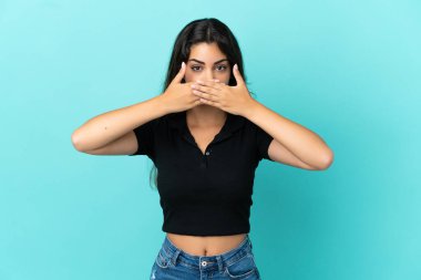 Mavi arka planda izole edilmiş genç beyaz kadın elleriyle ağzını kapatıyor.