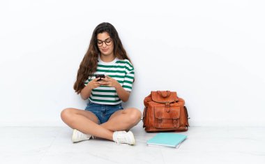 Genç bir kız öğrenci beyaz arka planda izole bir şekilde oturuyor ve cep telefonuyla bir mesaj gönderiyor.