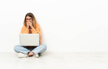 Dizüstü bilgisayarı olan genç bir kadın yerde oturuyor mutlu ve gülümsüyor elleriyle ağzını kapatıyor.