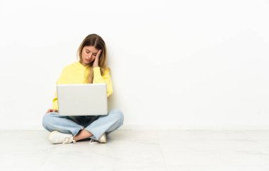 Dizüstü bilgisayarı olan genç bir kadın baş ağrısıyla yerde oturuyor.