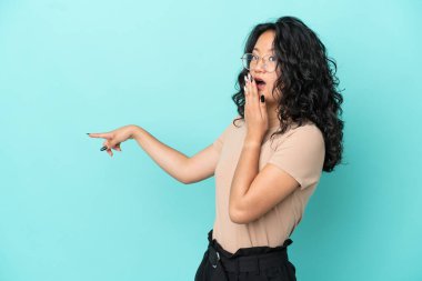Asyalı genç bir kadın mavi arka planda izole edilmiş, sürpriz bir ifadeyle işaret ederken...