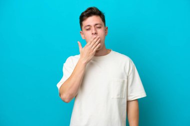 Genç ve yakışıklı Brezilyalı bir adam mavi arka planda izole edilmiş esniyor ve geniş bir ağzı eliyle kapatıyor.