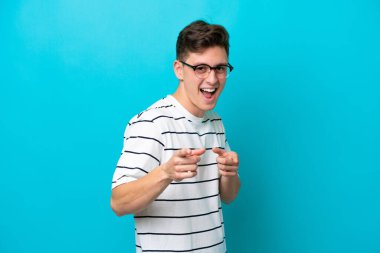 Genç ve yakışıklı Brezilyalı bir adam mavi arka planda izole edilmiş. Önü işaret ediyor ve gülümsüyor.