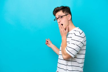 Genç, yakışıklı Brezilyalı bir adam mavi arka planda izole edilmiş, sürpriz bir ifadeyle işaret ederken...