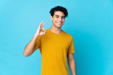 Genç Venezuelalı adam mavi arka planda izole edilmiş. Parmaklarıyla işaret veriyor.