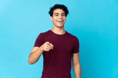 Genç Venezuelalı adam mavi arka planda izole edilmiş, şaşırmış ve ön tarafı işaret ediyor.