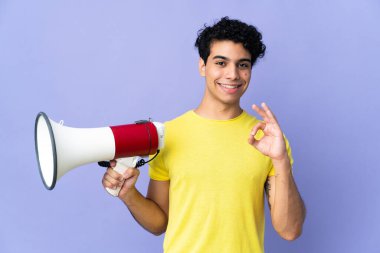 Genç Venezuelalı bir adam mor arka planda izole edilmiş bir şekilde elinde bir megafonla tamam işaretini parmaklarıyla gösteriyor.