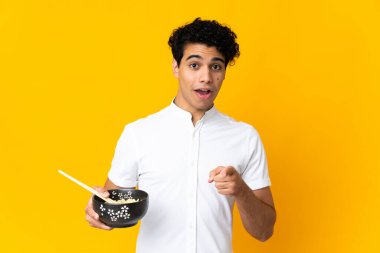 Genç Venezuelalı bir adam sarı arka planda izole edilmiş. Elinde yemek çubuklarıyla bir kase erişte tutarken şaşırmış ve ön tarafı işaret ediyor.