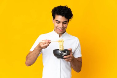 Genç Venezuelalı adam sarı arka planda izole edilmiş. Elinde çubuklu erişte kasesi var.