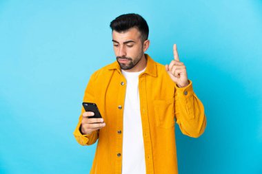 İzole edilmiş mavi arka plan üzerinde cep telefonu kullanan ve parmağını kaldıran beyaz bir adam.