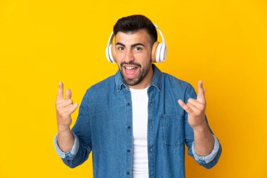 Kafkasyalı adam izole edilmiş sarı arka planda müzik dinliyor ve rock hareketleri yapıyor.