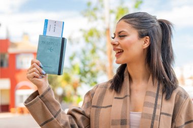 Genç ve güzel bir kadın elinde pasaportuyla dışarıda mutlu bir ifadeyle.