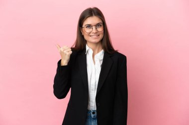 Pembe arka planda izole edilmiş genç beyaz kadın bir ürünü sunmak için kenarı işaret ediyor.