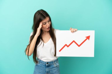 Mavi arka planda izole edilmiş genç beyaz bir kadın elinde büyüyen bir istatistik okuyla şaşırmış ifade ile bir işaret tutuyor.