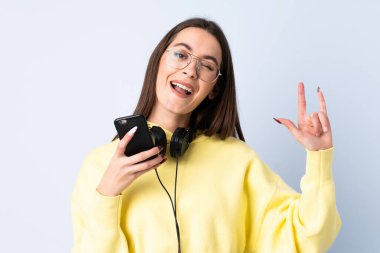 İzole edilmiş mavi arka planda gezici bir rock hareketi yapan genç bir kadın müzik dinliyor.