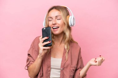 Pembe arka planda izole edilmiş genç bir Rus kadın cep telefonuyla müzik dinliyor ve şarkı söylüyor.